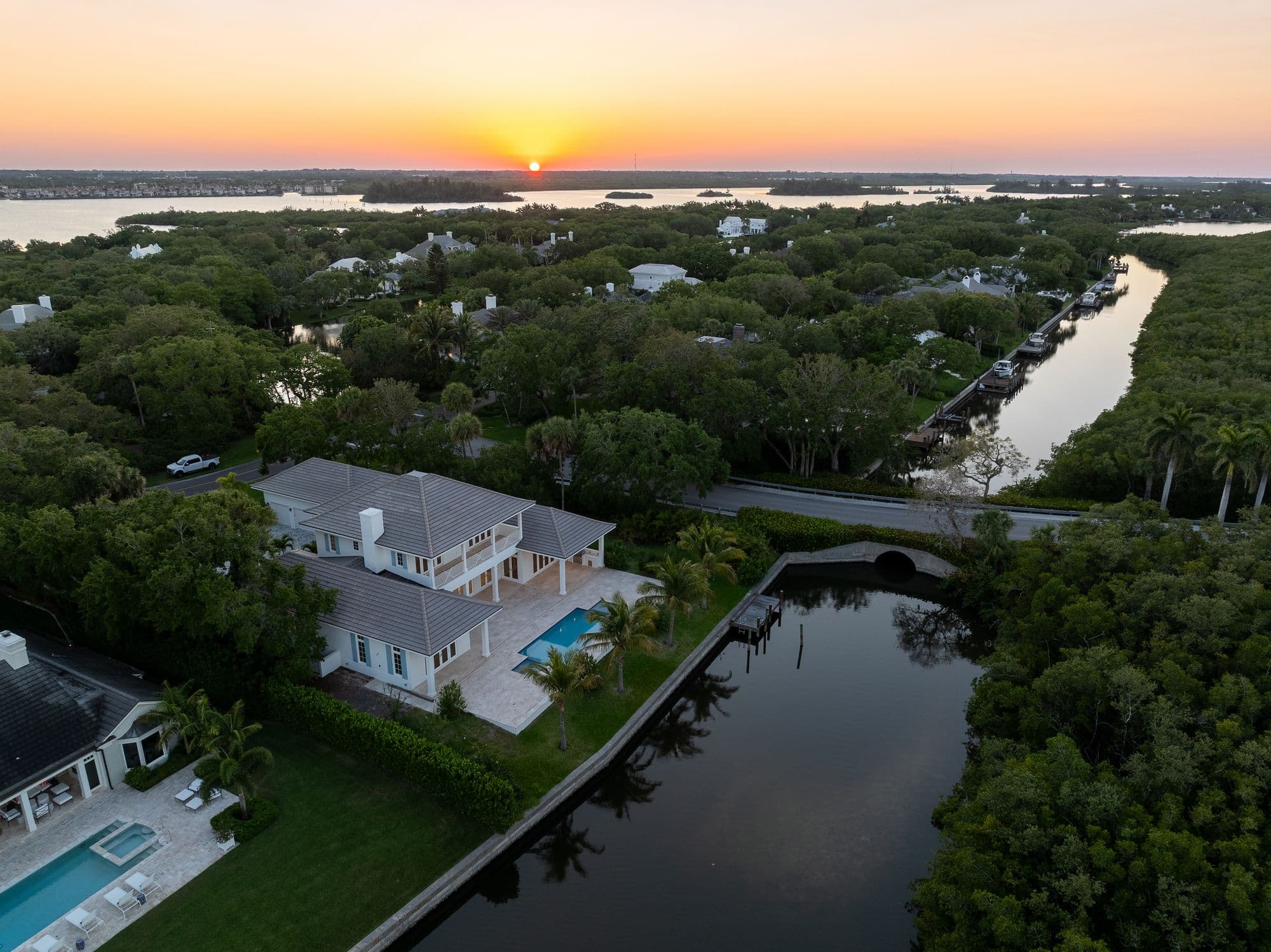 Aerial view of a large white house with a pool, next to a canal with a bridge, surrounded by dense trees at sunset.