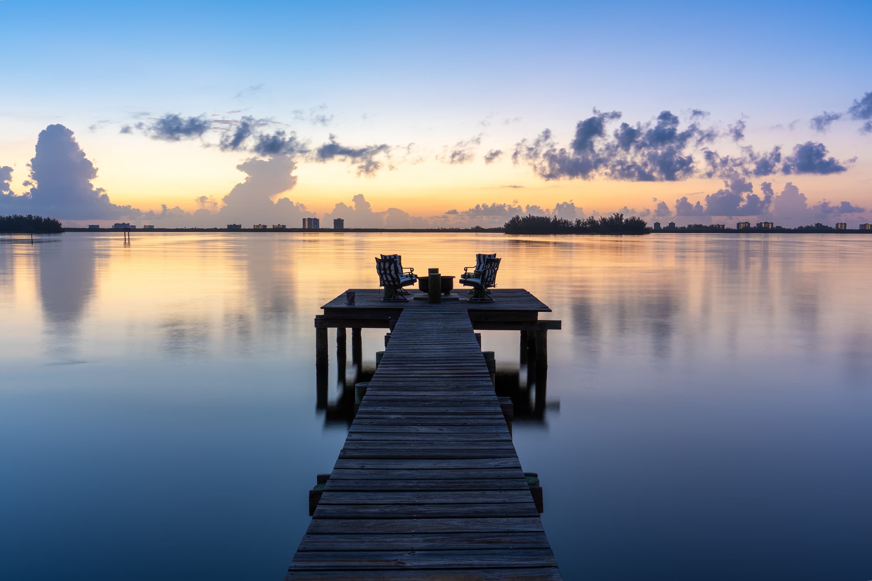 Dock leading into calm water with two chairs on an end platform against a colorful sunrise sky.