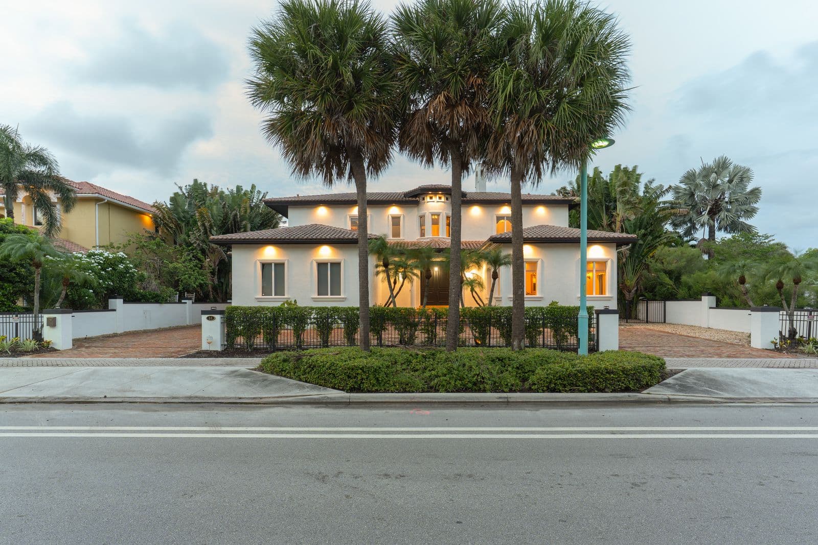 Exterior of a white two-story house with terracotta roof tiles, illuminated windows, and three tall palm trees.