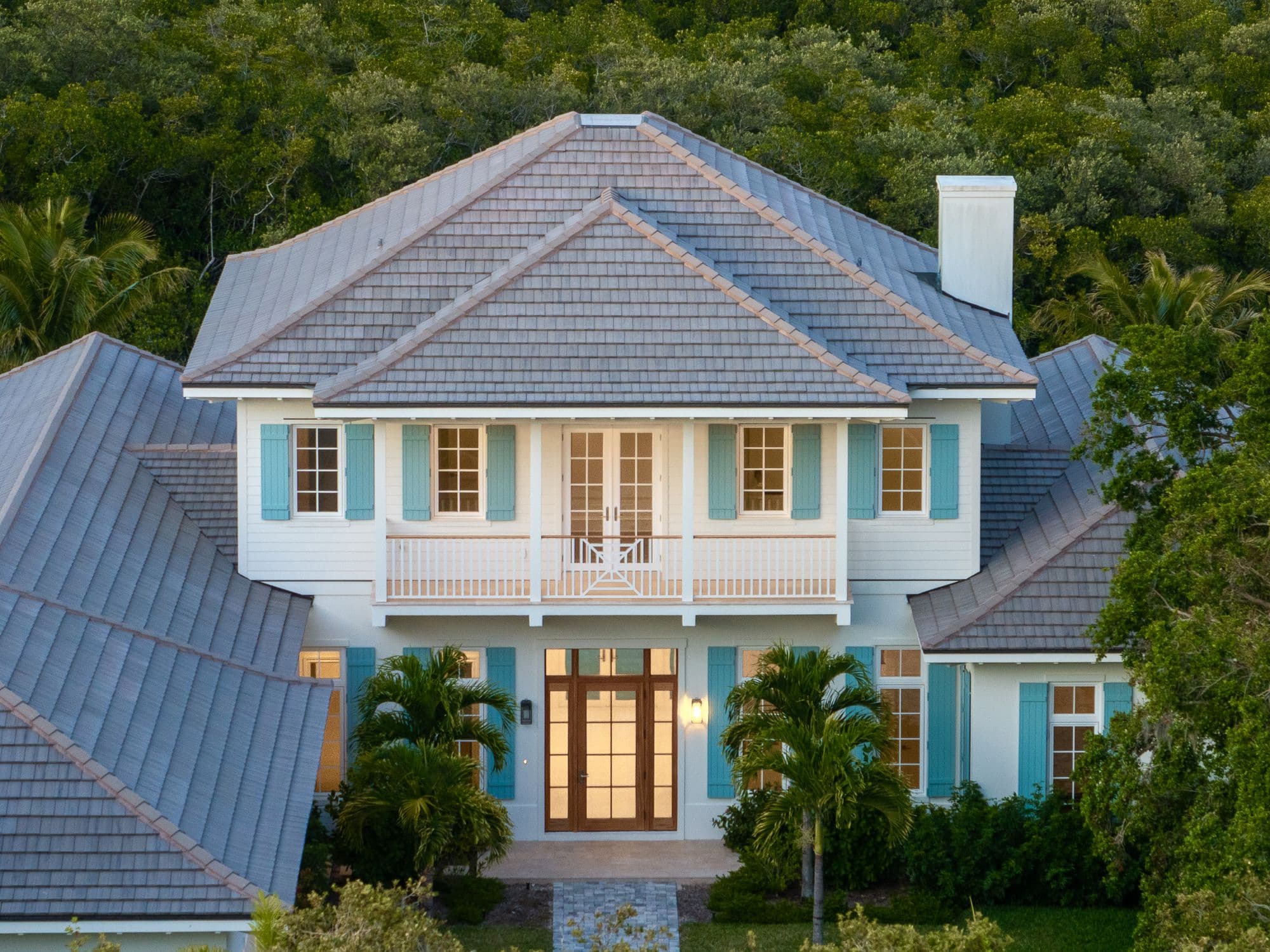White house exterior with grey shingle roof, blue shutters, a second-story balcony, and lit windows.