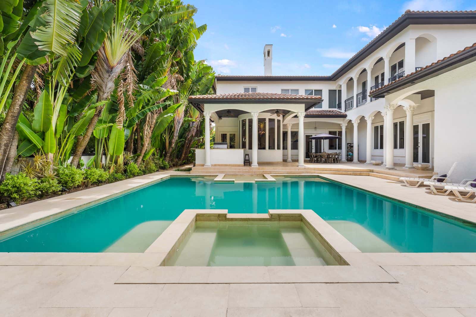 Large white house exterior with arched balconies, a turquoise pool, hot tub, and lush tropical trees.