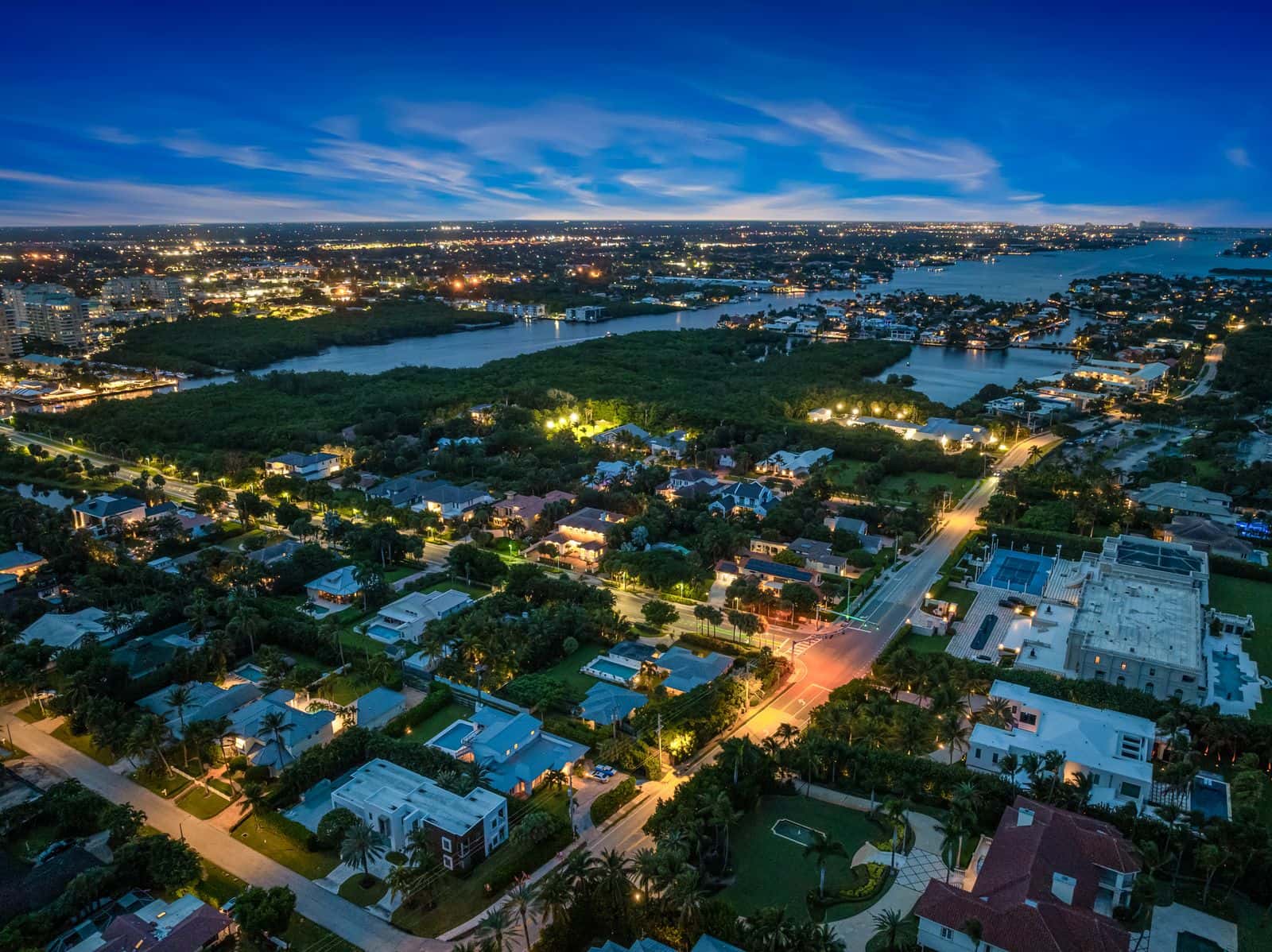 Aerial view of a coastal residential area at twilight, with illuminated homes, streets, and waterways.