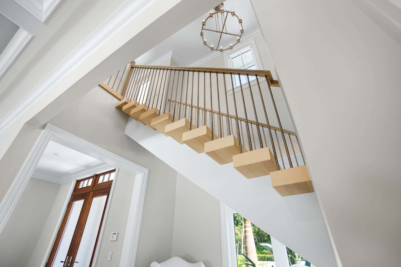Upward view of a modern floating staircase with light wood steps, metal balusters, and a chandelier.