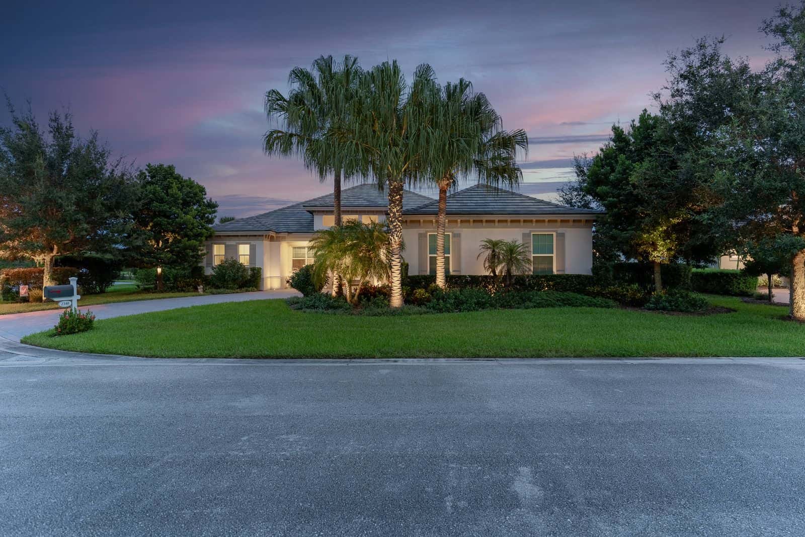 Exterior of a white single-story house at twilight, illuminated by exterior lights, with palm trees and green landscaping.