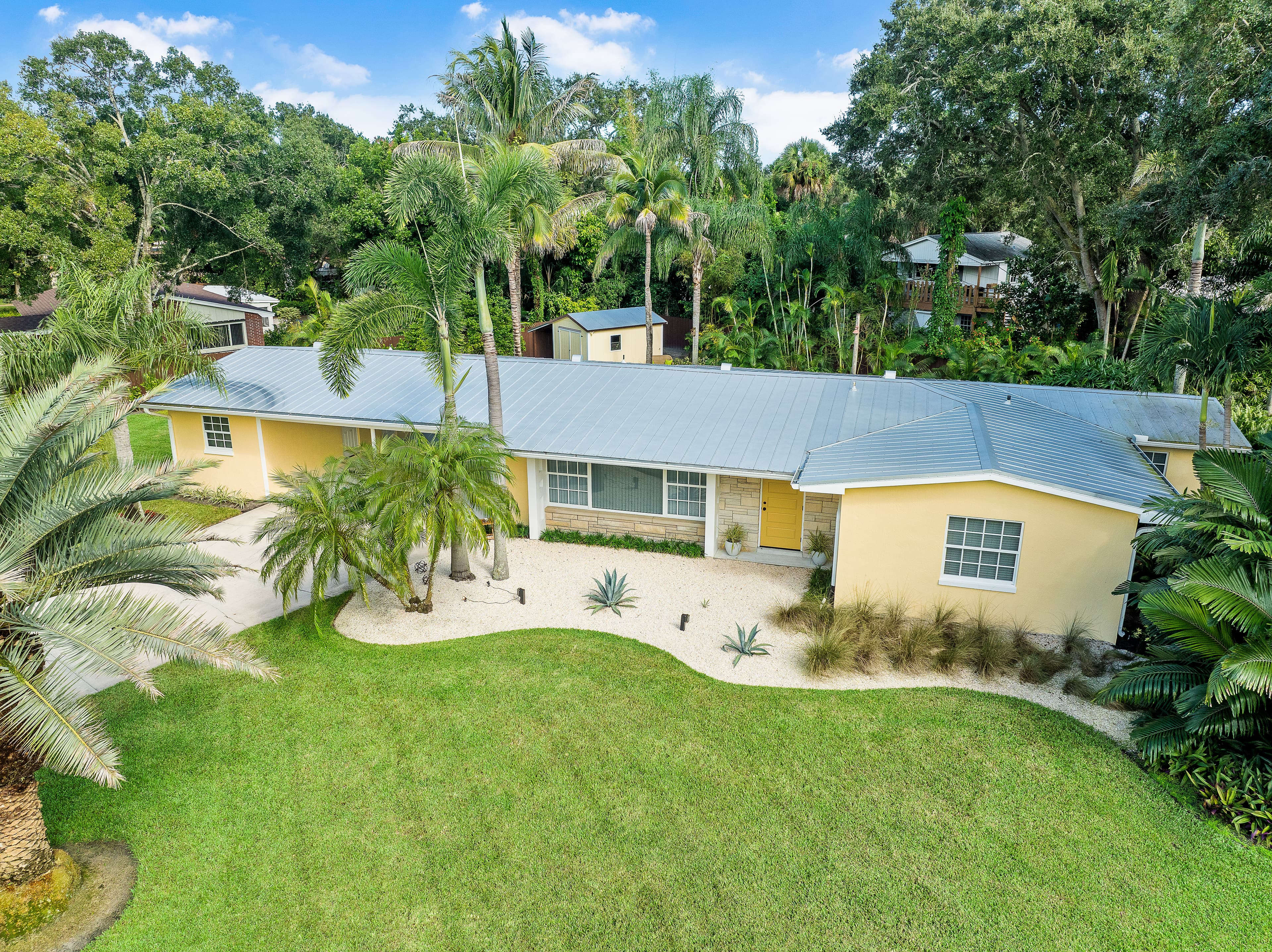 Exterior of a yellow house with a gray metal roof, tropical landscaping, and a green lawn.
